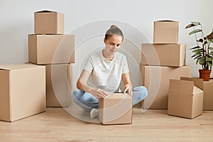 Indoor shot of attractive woman wearing white t shirt and jeans sitting on floor surrounded with cardboard boxes and unpacking