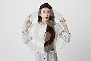 Indoor shot of beautiful brunette girl with long hair having excited, superstitious and naive look, keeping fingers