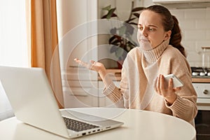 Indoor shot of beautiful Caucasian confused woman wearing beige jumper posing in kitchen and having video call on laptop, holding