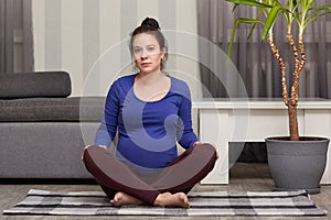 Indoor shot of beautiful European pregnant woman sits in zen posture, keeps legs crossed, has yoga exercises at home, looks