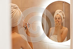 Indoor shot of beautiful woman being wrapped in white towel on her head smelling perfume while posing in bathroom, enjoying