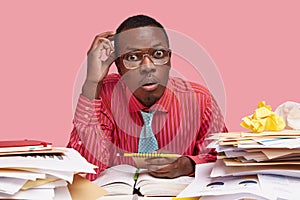 Indoor shot of black employee gazes with stupefaction, has widely opened eyes, wears spectacles, formal pink shirt photo