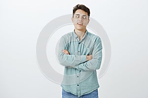 Indoor shot of bored childish handsome guy in casual shirt, holding hands crossed on chest and squinting, standing