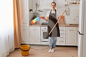 Indoor shot of bored young woman housekeeper holding brush and ppduster, washing her kitchen, wearing brown apron, jeans and white