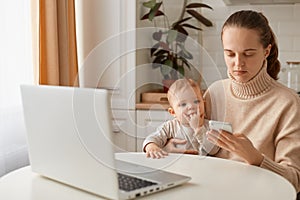 Indoor shot of Caucasian dark haired woman wearing beige sweater sitting at table with baby girl in hands and using cell phone,