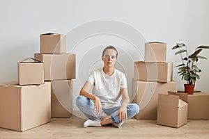 Indoor shot of Caucasian woman wearing white t shirt sitting with crossed legs on floor surrounded with cardboard boxes with