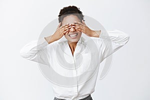 Indoor shot of charming carefree african-american female employer with white shirt, covering eyes with palms, being