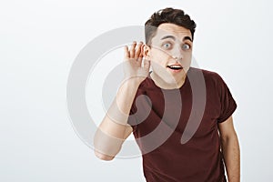 Indoor shot of charming european man in casual outfit, bending towards camera and holding hand near eay, overhearing