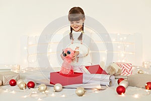 Indoor shot of charming little girl wearing white sweater holding soft toy, looking at her New year gift with smile, sitting on
