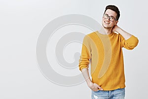 Indoor shot of charming nice and handsome young guy in glasses and yellow sweater touching back of neck shy and unsure