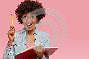 Indoor shot of cheerful dark skinned student keeps pencil in hand, feels happy as gets good idea for writing