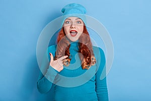 Indoor shot of cheerful smiling young Caucasian woman with curls pointing index finger on her self, keeps mouth opened, posing