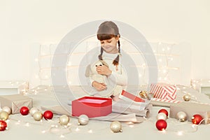 Indoor shot of child sitting on bed on oft blanket with a gift in her hands, opening box with Christmas present, posing in bedroom