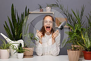 Indoor shot of confused caucasian adorable brown-haired woman florist wearing white shirt sitting in flower store with garden