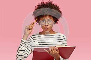 Indoor shot of confused indecisive woman with black skin, curly hair, scratches head with pencil, dressed in striped