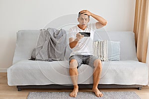 Indoor shot of confused man wearing white t shirt and jeans shirt sitting on sofa at home, holding cell phone in hands, playing