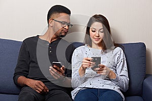 Indoor shot of couple enjoying media content in smart phone sitting on comfortable couch at home, holding mobile phones, dark