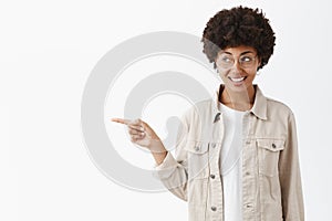 Indoor shot of creative and stylish boyish woman in glasses and cool beige shirt pointing and gazing left while
