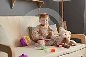 Indoor shot of cute baby girl playing with plush toys and plastic sorter, sitting on sofa alone, little kid with educational toy.