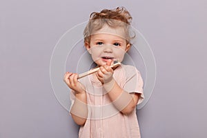 Indoor shot of cute blonde wavy haired toddler baby girl brushing her teeth, looking at camera, playing with tooth, standing