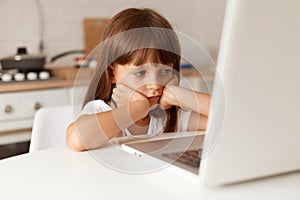 Indoor shot of cute bored dark haired female child sitting at table, looking at notebook screen with boring facial expression,