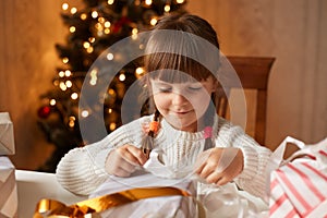 Indoor shot of cute charming little girl wearing white sweater packing presents for her parents and friends, smiling happily while