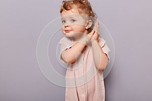 Indoor shot of cute curios little toddler caucasian girl posing isolated over gray background, baby with blond curly hair keeps