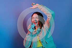 Indoor shot of cute positive young girl wearing casual jacket standing isolated over blue neon light background, holding smart