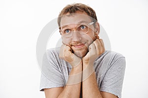 Indoor shot of cute and tender adult guy in glasses and grey t-shirt smiling with delight leaning head on palms and