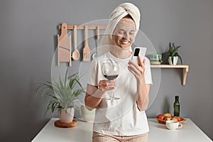 Indoor shot of delighted attractive woman with cosmetic mask in white towel wrapped around head drinking wine in kitchen, doing