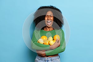 Indoor shot of emotional curly girl holding fresh oranges and lemons fruits, keeps mouth widely opened, dressed in