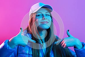 Indoor shot of emotional teenage lady pointing fingers at herself while standing isolated over neon pink background, looking at