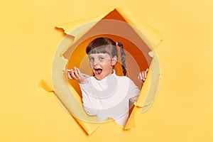 Indoor shot of excited amazed little girl with pigtail breaking through yellow paper and holding cell phone, recording voice