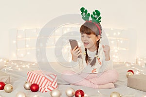 Indoor shot of excited little adorable girl wearing white casual sweater sitting on bed with cell phone in hands, clenched fists,