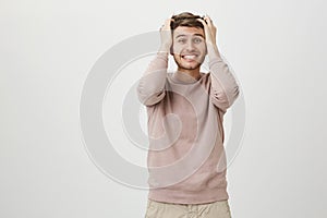 Indoor shot of excited and overwhelmed guy smiling broadly and looking with widened eyes while holding hands on hair and