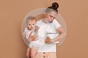 Indoor shot of exhausted shocked woman wearing white casual style shirt holding in arms toddler daughter, looks at the spoiled