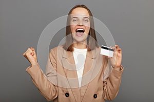 Indoor shot of exited extremely happy brown-haired woman wearing jacket holding credit card isolated over gray background,