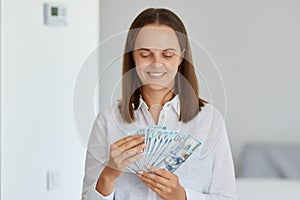 Indoor shot of extremely happy female wearing white shirt holding fan of banknotes in hands, dreaming to buy lots things, winning