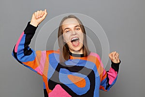 Indoor shot of extremely happy joyful woman with brown hair wearing colorful jumper, clenched fists, dancing, rejoicing her