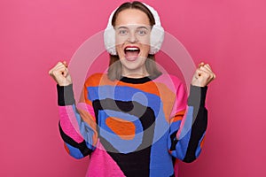 Indoor shot of extremely happy young adult woman wearing warm sweater and fur earmuffs clenched fists, celebrating her success,