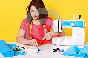 Indoor shot f concentraed woman dressmaker sewing in her studio, sitting with opened mouth, ready to sew buttons, has confident