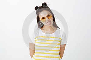 Indoor shot of fashionable picky and moody girlfriend in stylish outfit and glasses with hair buns, sticking out tongue photo