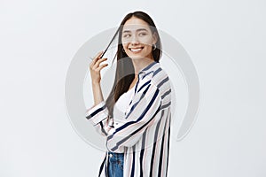 Indoor shot of feminine flirty fashionable woman in striped blouse, turning right while playing with hair strand and