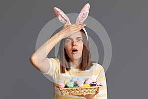 Indoor shot of frighten astonished woman wearing rabbit ears holding colorful Easter eggs isolated on gray background, female