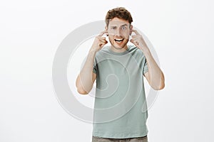 Indoor shot of funny caucasian boyfriend in casual t-shirt, making faces, covering hearing with index fingers in ears