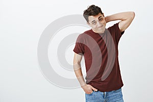 Indoor shot of guilty embarrassed european male model in casual red t-shirt, scratching back of neck and looking from photo