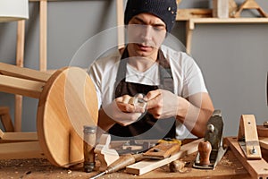 Indoor shot of handsome carpenter making a handmade wooden toy in a home workshop, enjoying wood carving, posing in his joinery,