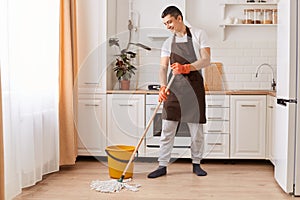 Indoor shot of handsome man brown apron mopping the floor in the kitchen, expressing positive emotions, doing household work,