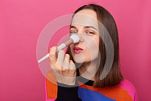 Indoor shot of happy attractive beautiful woman doing daily makeup, applying cosmetic on her hose, looking at camera with happy