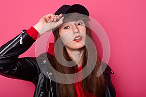 Indoor shot of happy Caucasian young teenager girl in red hoodie, leather jacket, stands with cheeky facial expression, keeps hand